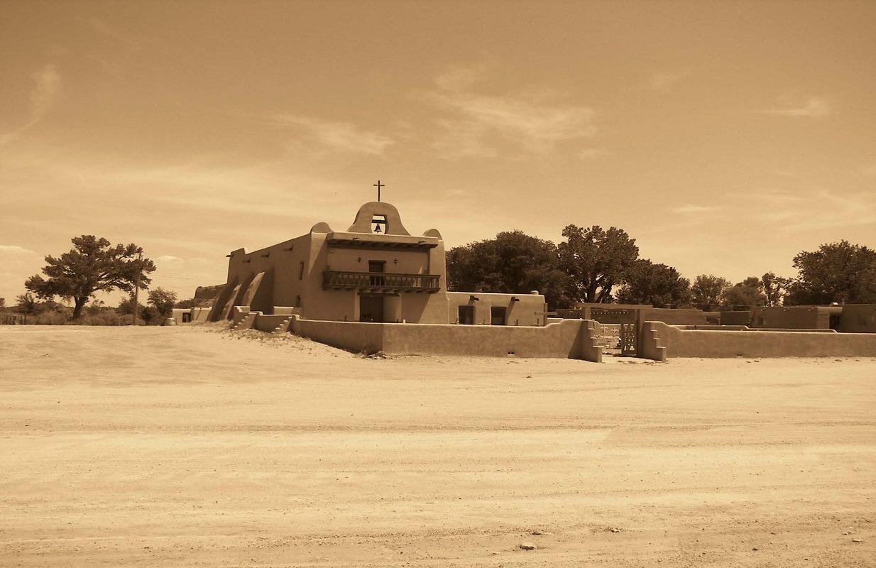 Historic adobe building with a cross and balcony, surrounded by sandy terrain and trees, reflecting cultural architecture.