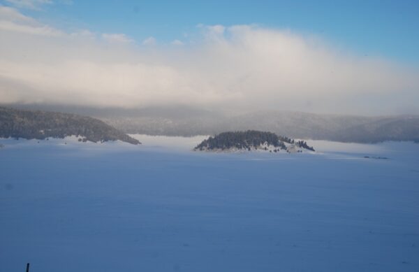 Serene snowy landscape featuring a frozen lake surrounded by lightly wooded hills under a partly cloudy blue sky