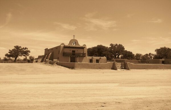 Historic adobe building with a cross and balcony, surrounded by sandy terrain and trees, reflecting cultural architecture.